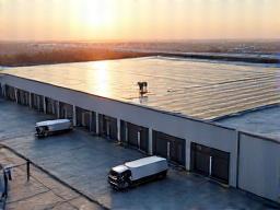 Vast warehouse complex with an expansive rooftop solar array and several electric trucks charging at designated stations, showcasing integrated green logistics.